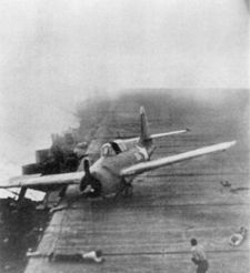 A Hornet Wildcat that just landed minutes earlier skids across Enterprise’s flight deck as the carrier maneuvers violently during Junyo’s dive bomber attack. Two crewmen are taking defensive postures on the deck as smoke from earlier bomb hits swirls around them.